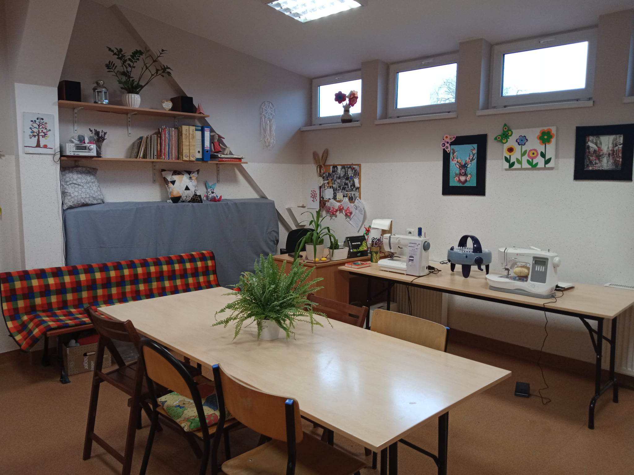 zdjęcie pracowni krawiecko-dziewiarskiej, stół z kwiatem na środku, maszyny do szycia, deska do prasowania, ławka do odpoczynku